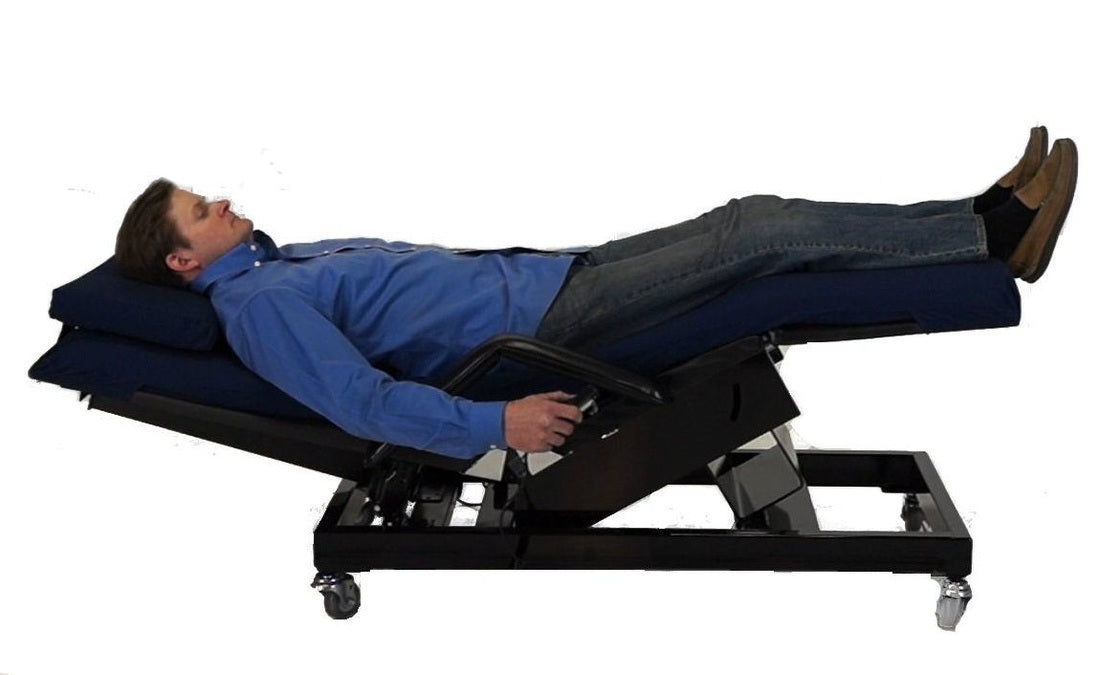 A man reclining on a black motorized adjustable massage table with a dark blue padded headrest and footrest, shown against a plain white background. Zero Gravity Chair 5 Special Needs Computers.ca