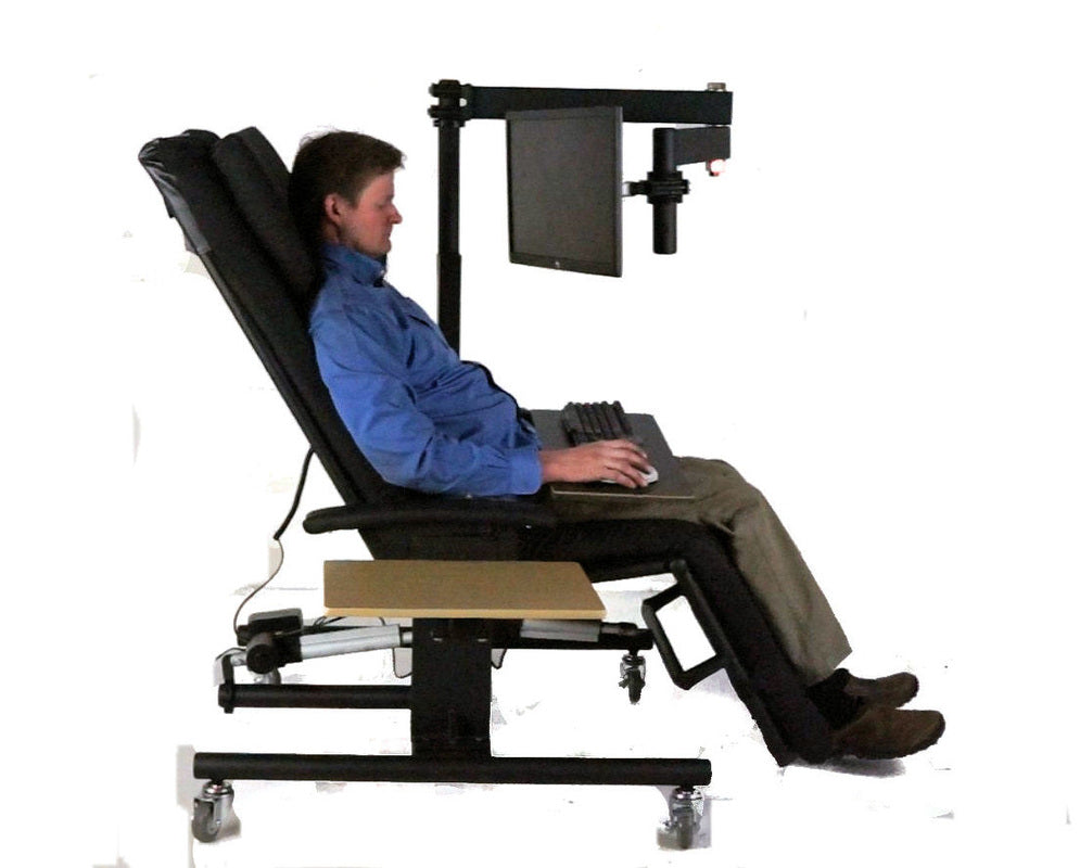 A man seated in a black adjustable medical-style reclining chair with a mounted monitor, keyboard, and footrest, photographed against a plain white studio background. Zero Gravity Chair 1a Special Needs Computers.