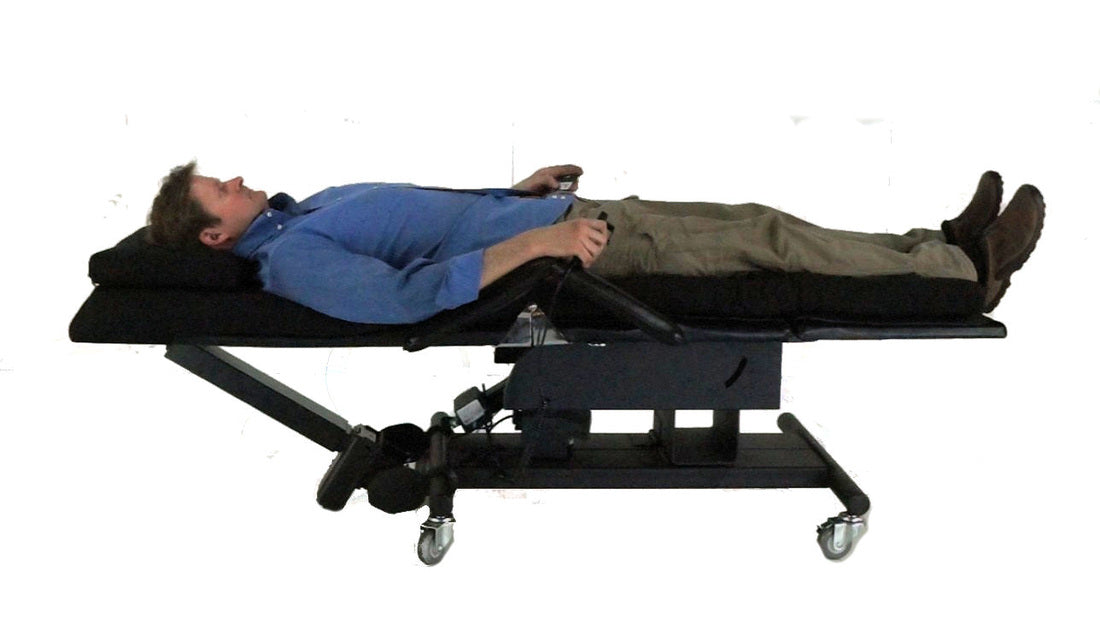 A man lies on a black adjustable medical-style table or gurney with wheels, photographed against a plain white background. Zero Gravity Chair 3a