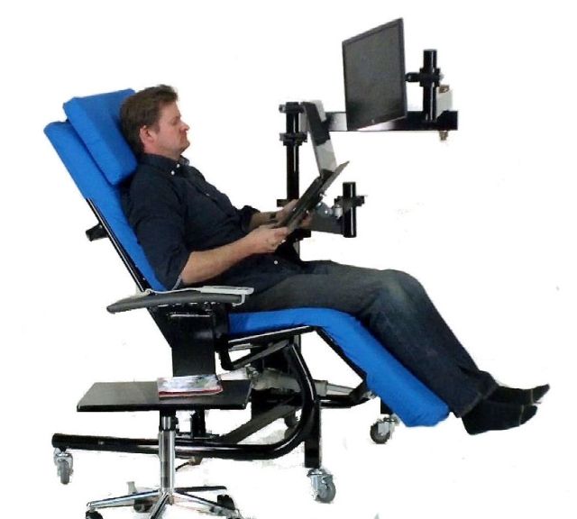 A man reclining in a blue and black ergonomic office chair with a built-in monitor arm and screen, holding a tablet, on a white background. Zero Gravity Workstation 1c. Special Needs Computers Canada.