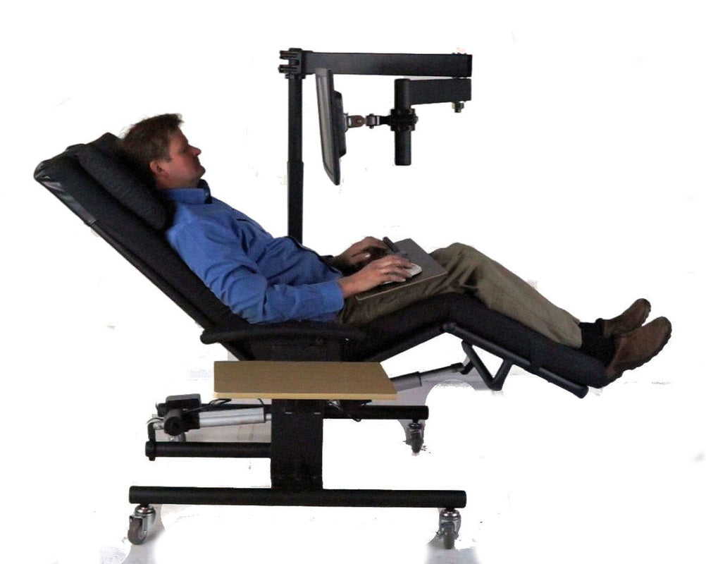 A reclining man using a computer on a black adjustable ergonomic chair with a mounted monitor and keyboard tray, shown against a plain white background. Zero Gravity Chair 1a Special Needs Computers.