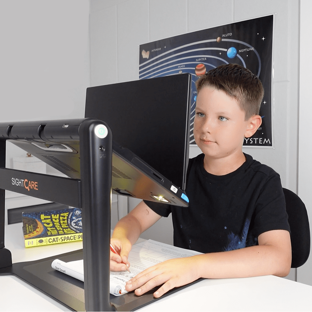 A boy sitting at a desk using a black SightCare ergonomic monitor stand with a computer monitor and keyboard. A 'CAT SPACE PROGRAM' book is propped up on the stand, and a space-themed poster is on the wall behind him. Special Needs Computers.