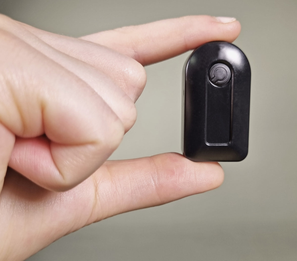 A small black personal alarm or key finder device held between two fingers to show its compact size, against a neutral gray background.