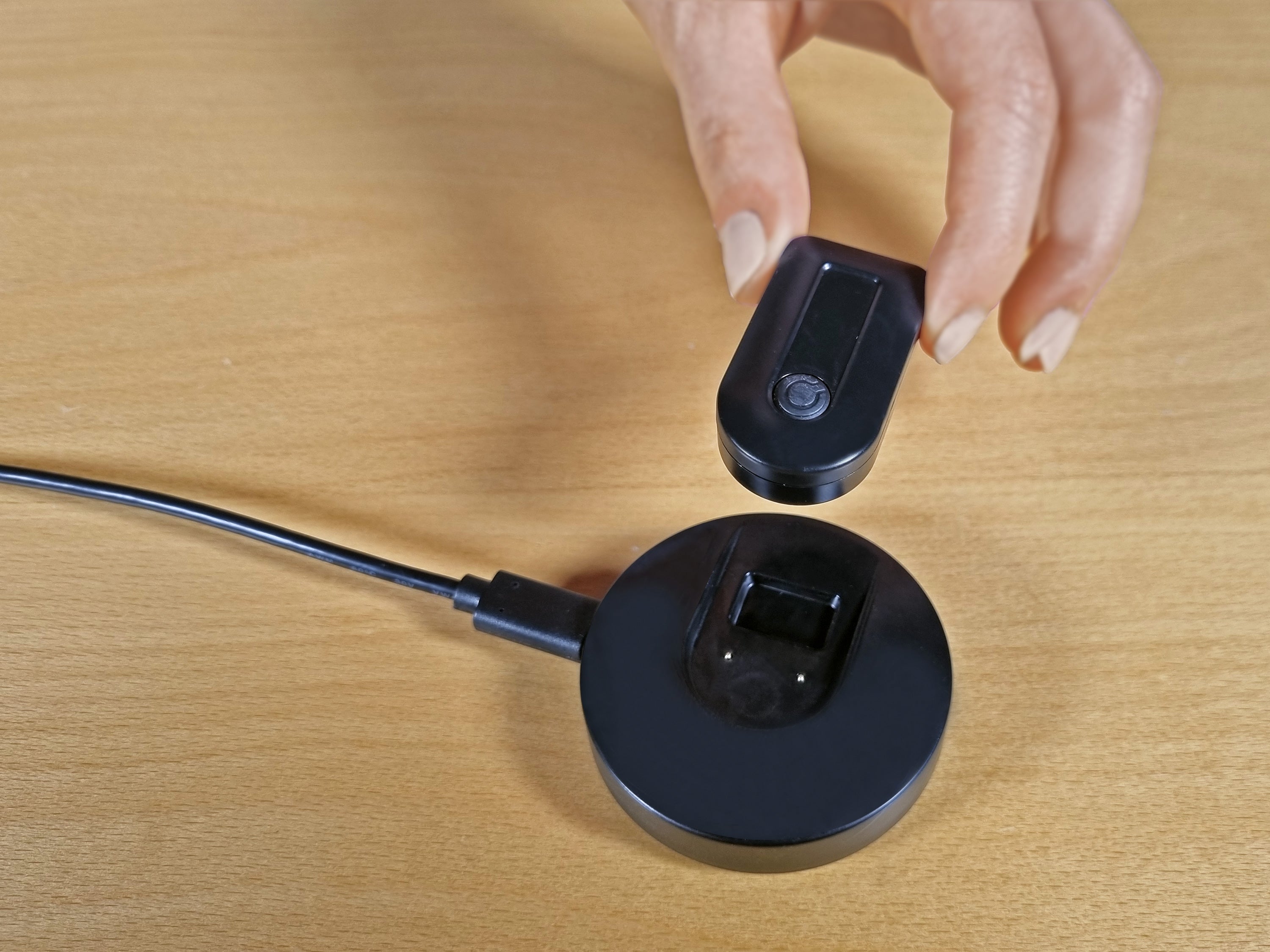 A close-up of a person's hand placing a small black rectangular electronic device onto a matching round black charging dock with a cable, set on a light wood tabletop.
