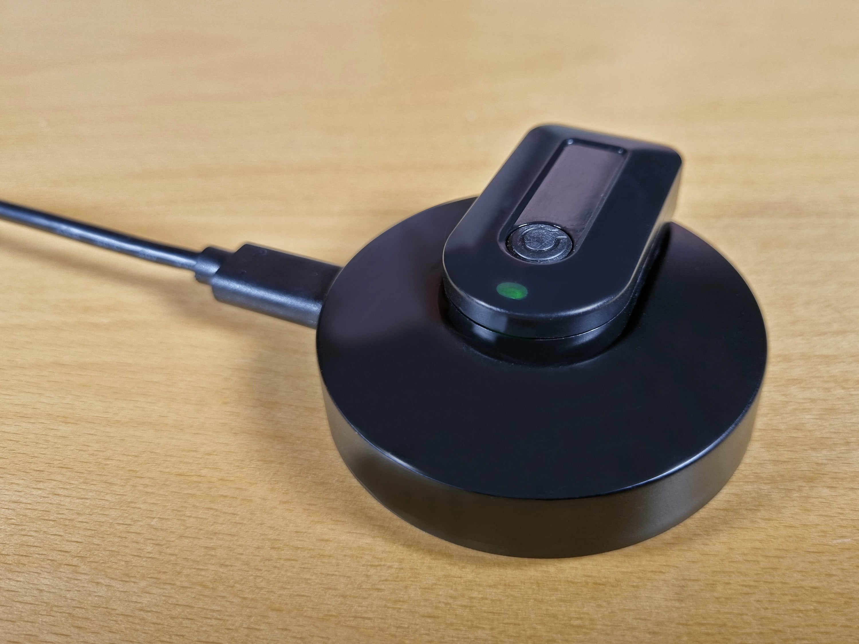 A black circular wireless charging pad with a matching charging dock on top, connected to a cable, resting on a light wood surface.