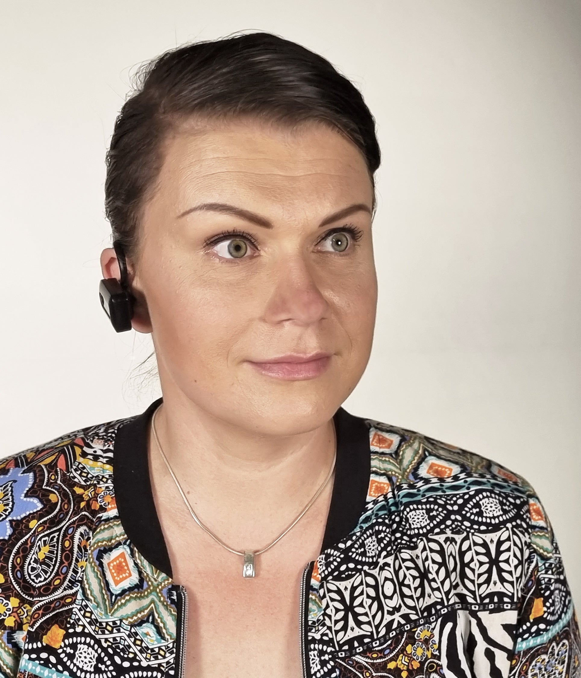 A woman with dark hair pulled back, wearing a black wireless earbud and a patterned black jacket with orange, blue, and white details, against a plain light background.