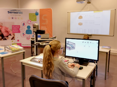 A young girl with a ponytail sits at a desk using a computer in a classroom or workshop setting, with a Sensotec display board and a projector screen visible in the background. Special Needs Computers