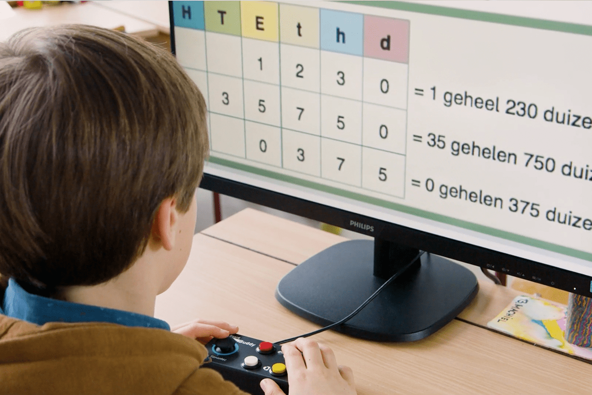 A child in a brown jacket sits at a desk, looking at a computer monitor showing a Dutch multiplication learning game. The child is holding a black game controller. Special Needs Computers