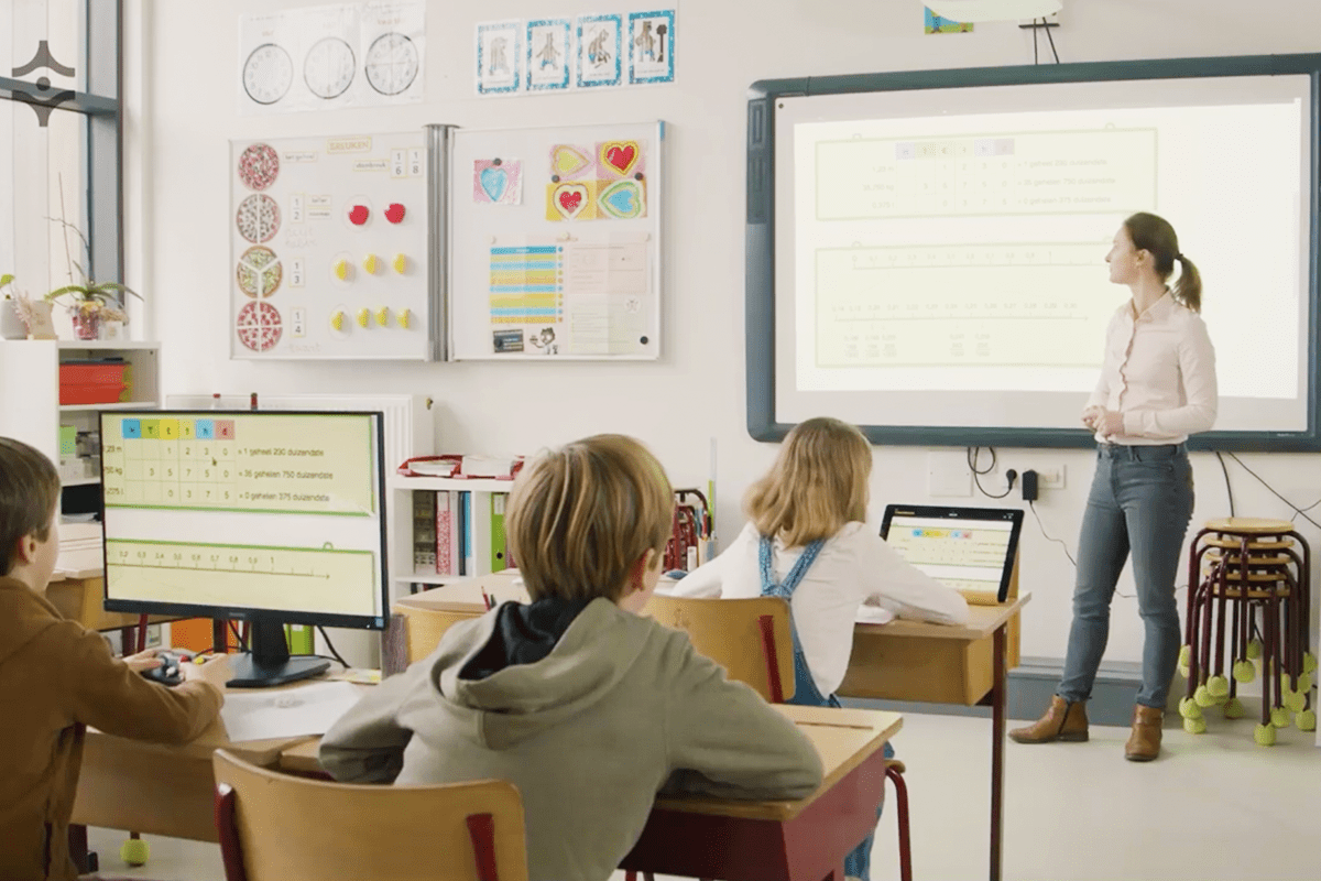 A classroom scene with a teacher standing at a whiteboard while students sit at desks using computers. The room has educational posters, shelves, and a bright whiteboard showing lesson content. Special Needs Computers
