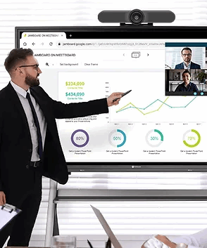 A man in a black suit stands in front of a large interactive whiteboard screen, pointing at charts and graphs while a webcam on a monitor shows a virtual meeting with two other men. Special Needs Computers
