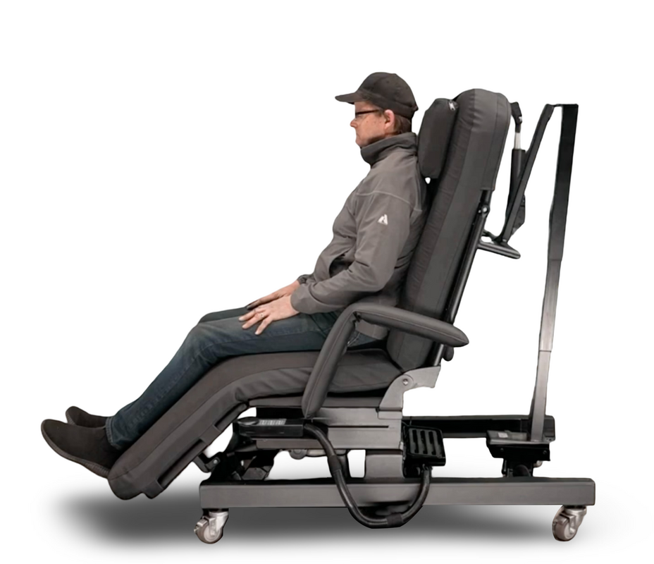 A man wearing a cap and glasses sits in a dark gray adjustable medical-style reclining chair with a headrest, armrests, and casters on a white studio background. Zero Gravity Chair 4 Special Needs Computers.