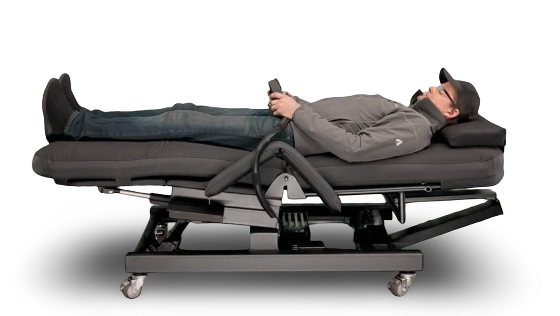A man lies on a dark gray adjustable electric massage table with wheels, wearing a gray long-sleeve shirt, blue jeans, and a black cap, while holding a smartphone. The table has a black metal frame and control panel. Zero Gravity Chair 4 Special Needs Computers.