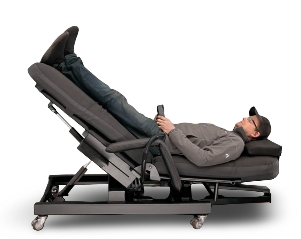 A man reclining on a dark gray adjustable power chaise lounge chair with his legs raised, holding a smartphone, shown against a plain white background. Zero Gravity Chair 4 Special Needs Computers.