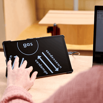 A hand holding a black slate board with white text and arrows, connected to a computer monitor on a wooden desk in a classroom setting.  Special Needs Computers