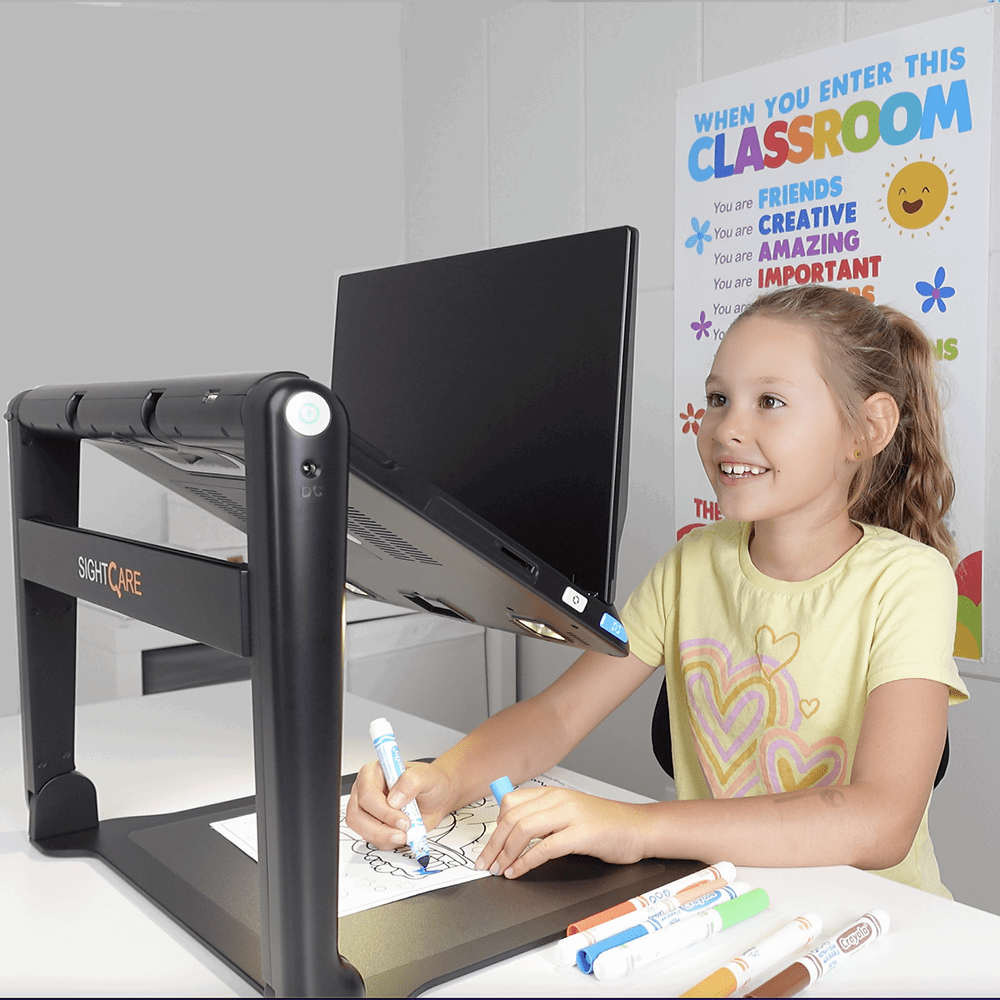 A child using a black SightCare digital tablet on a white desk, with a marker in hand. A SightCare brand sign is visible on the left side of the stand, and a colorful classroom poster is in the background. Special Needs Computers.