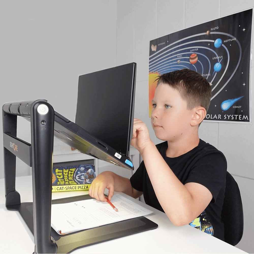 A young boy wearing a black t-shirt sits at a white desk using a black ergonomic monitor stand with an angled computer monitor. A science poster of the solar system is visible on the wall behind him.  Special Needs Computers.