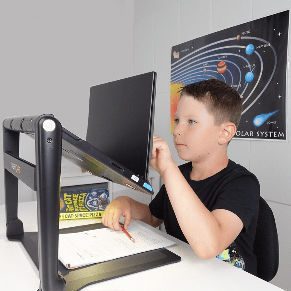 A young boy wearing a black t-shirt sits at a white desk using a black ergonomic monitor stand with an angled computer monitor. A science poster of the solar system is visible on the wall behind him.  Special Needs Computers.