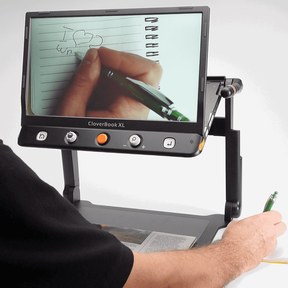 A CloverBook XL digital eye tracker and drawing tablet setup shown in use, with a person holding a green pen and looking at a screen that displays a hand writing in a notebook. The black device has control buttons on the front and stands on a base with an arm extending downward. CloverBook XL Special Needs Computers in Canada.