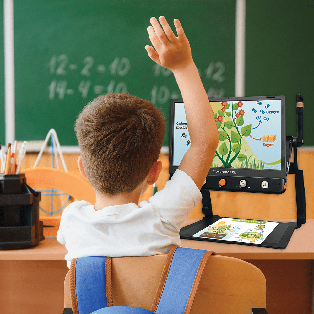 A child sitting at a desk in a classroom raises their hand while using a handheld educational tablet with a digital plant diagram on screen. The tablet is mounted on a stand on the desk, with a green chalkboard in the background. CloverBook XL Special Needs Computers in Canada.