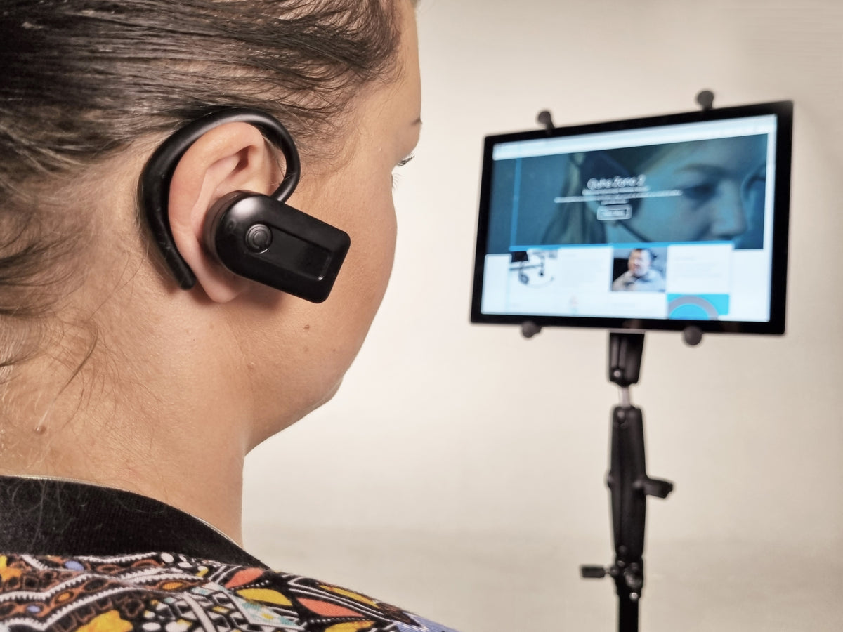 A woman in profile wearing a black behind-the-ear wireless earpiece, with a tablet on a stand displaying a video call interface in the background.