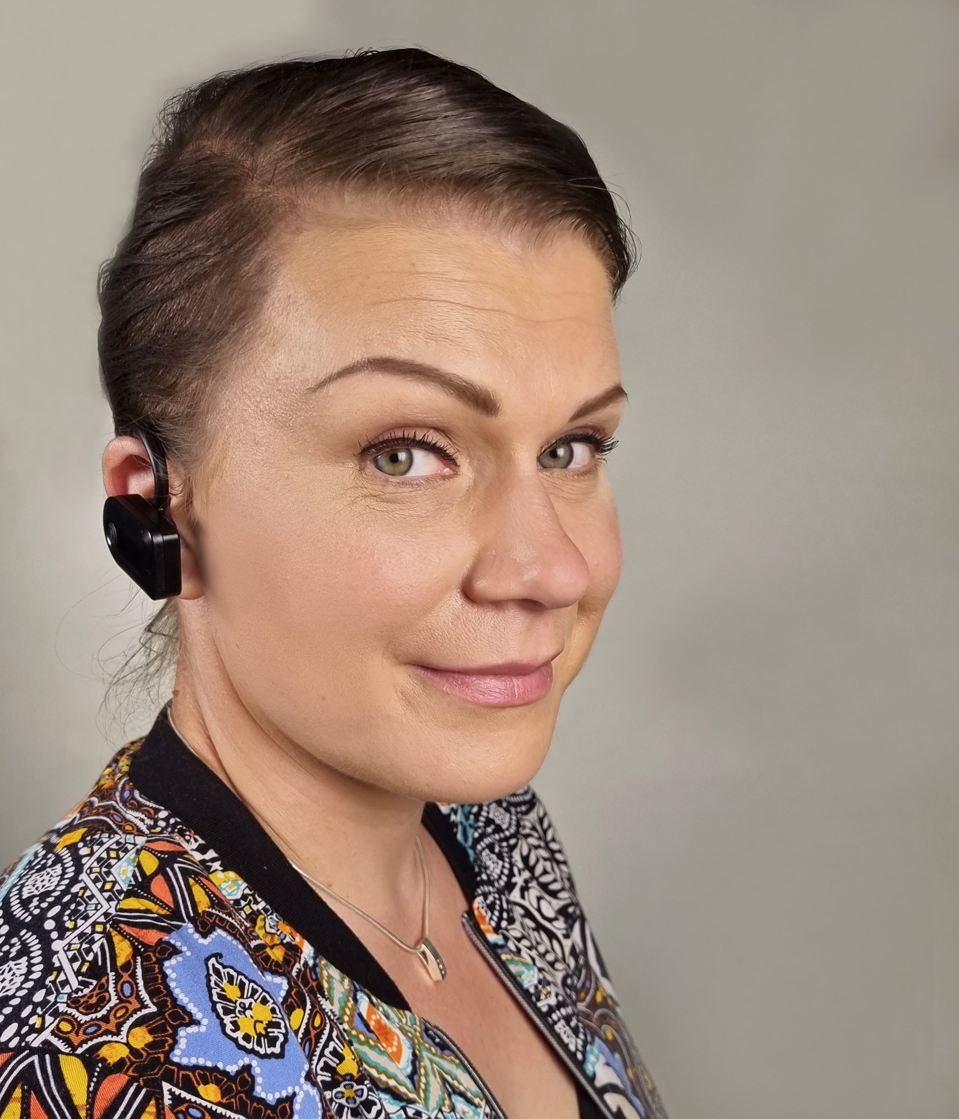 A woman with her hair pulled back wears a black in-ear earbud. She is wearing a colorful patterned jacket with black trim, set against a plain gray studio background.