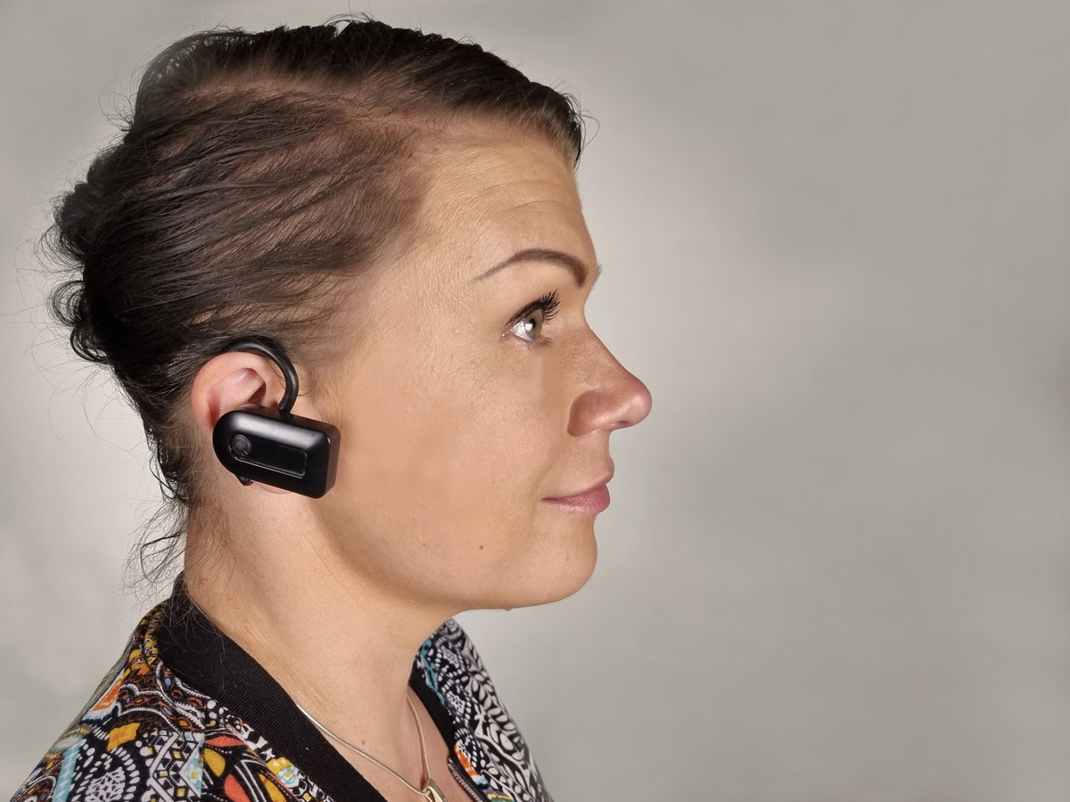 A woman in profile wearing a black wireless earbud over her ear, shown against a neutral gray background.