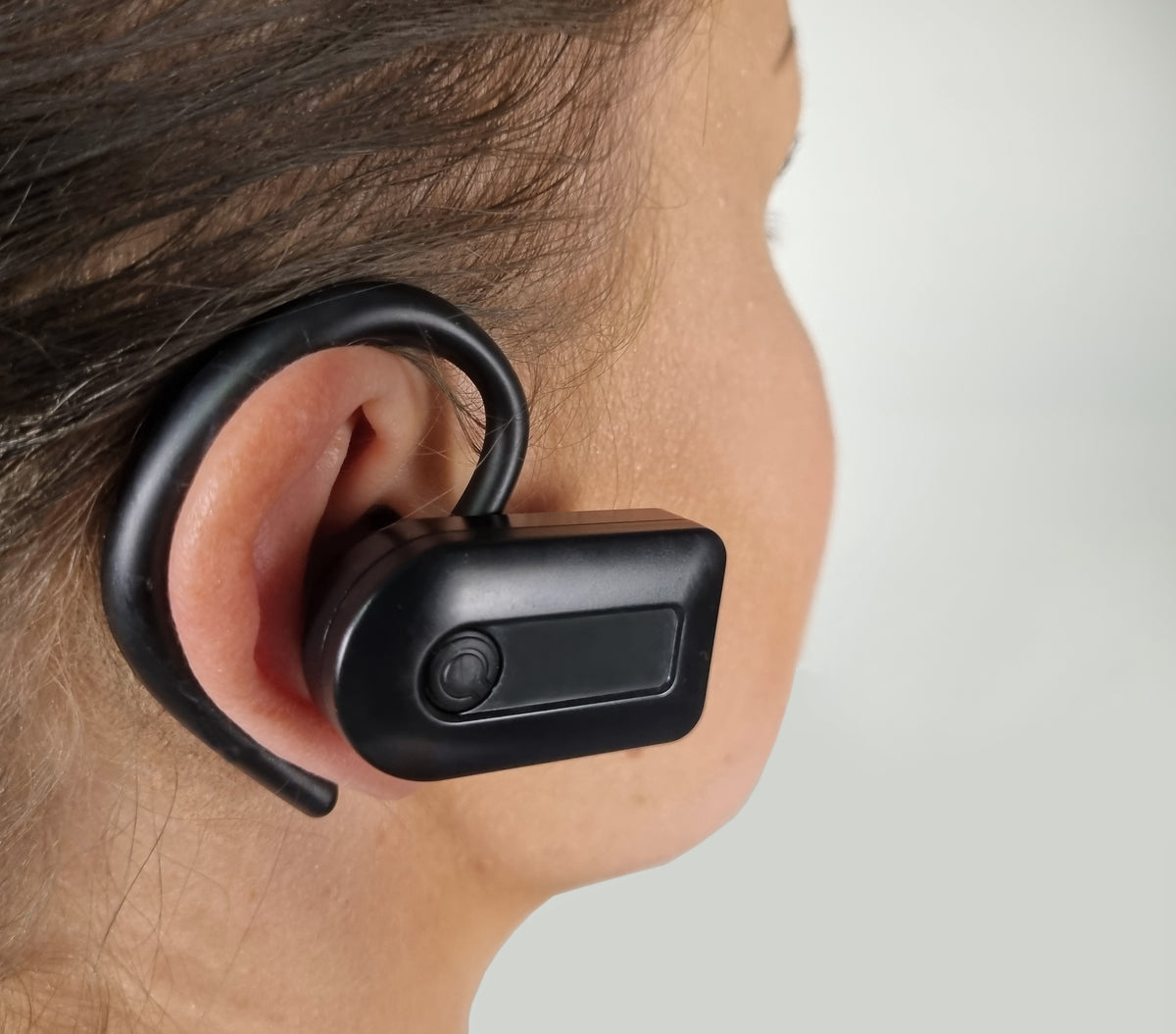 A close-up side profile of a person wearing a black wireless earbud with an ear hook against a light gray background.