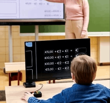 A child at a desk in a classroom using a black interactive whiteboard computer with a mouse, while a teacher stands near a larger screen in the background. Special Needs Computers