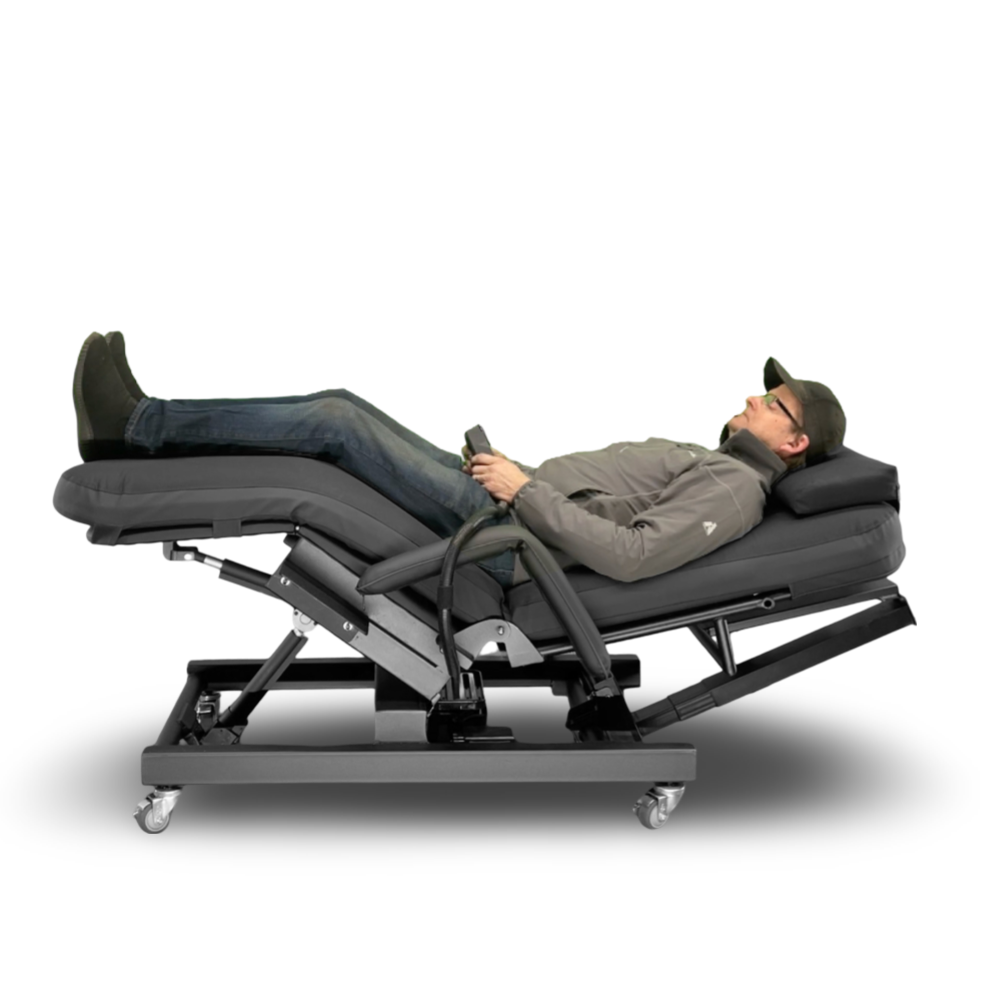 A man reclining on a motorized adjustable massage chair with his legs extended, holding a remote control. The chair is upholstered in dark gray fabric with a black metal frame on casters, shown against a plain white background. Zero Gravity Chair 4 Special Needs Computers.