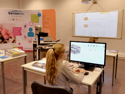 A young girl with a ponytail sits at a desk using a computer in a classroom or workshop setting, with a Sensotec display board and a projector screen visible in the background. Special Needs Computers