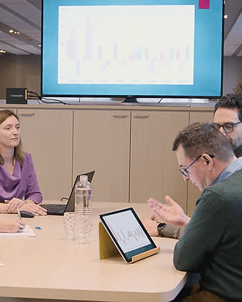 A group of three business professionals gathered around a table in a meeting room, with a tablet showing a graph in the foreground and a large screen behind them displaying a colorful business chart.  Special Needs Computers