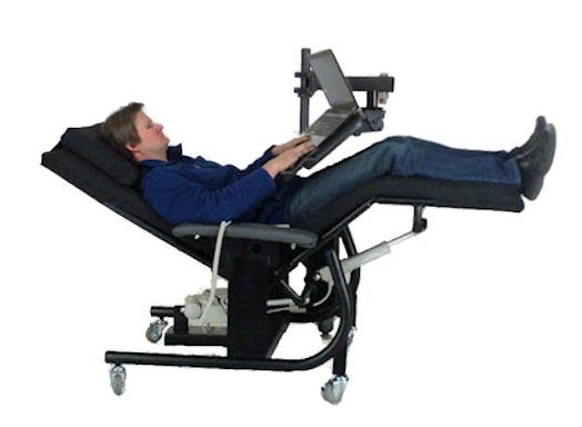 A man reclining in a black adjustable medical-style chair with a built-in laptop tray table, shown on a plain white background. Zero Gravity Workstation 1b. Special Needs Computers.