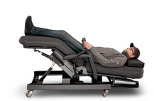 An electric massage table with a reclining patient, shown on a plain white background. The table has a dark gray frame and base with wheels, and the massage bed is upholstered in dark gray and black fabric. Zero Gravity Chair 4 Special Needs Computers.