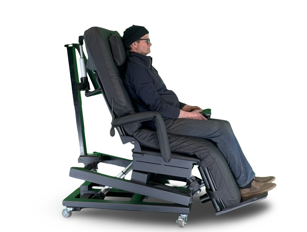 A man wearing a black beanie and glasses sits in a dark gray adjustable motorized recliner chair with green structural supports and wheels, shown from a side profile against a white background.  Zero Gravity Chair 4-Lift. Special Needs Computers.ca